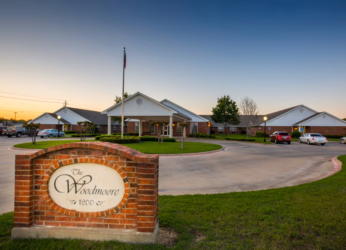 Brick entrance sign reading "The Woodmoore 1200" in front of a single-story brick senior living building with a covered entrance, flagpole, circular driveway, and parked cars at dusk.
