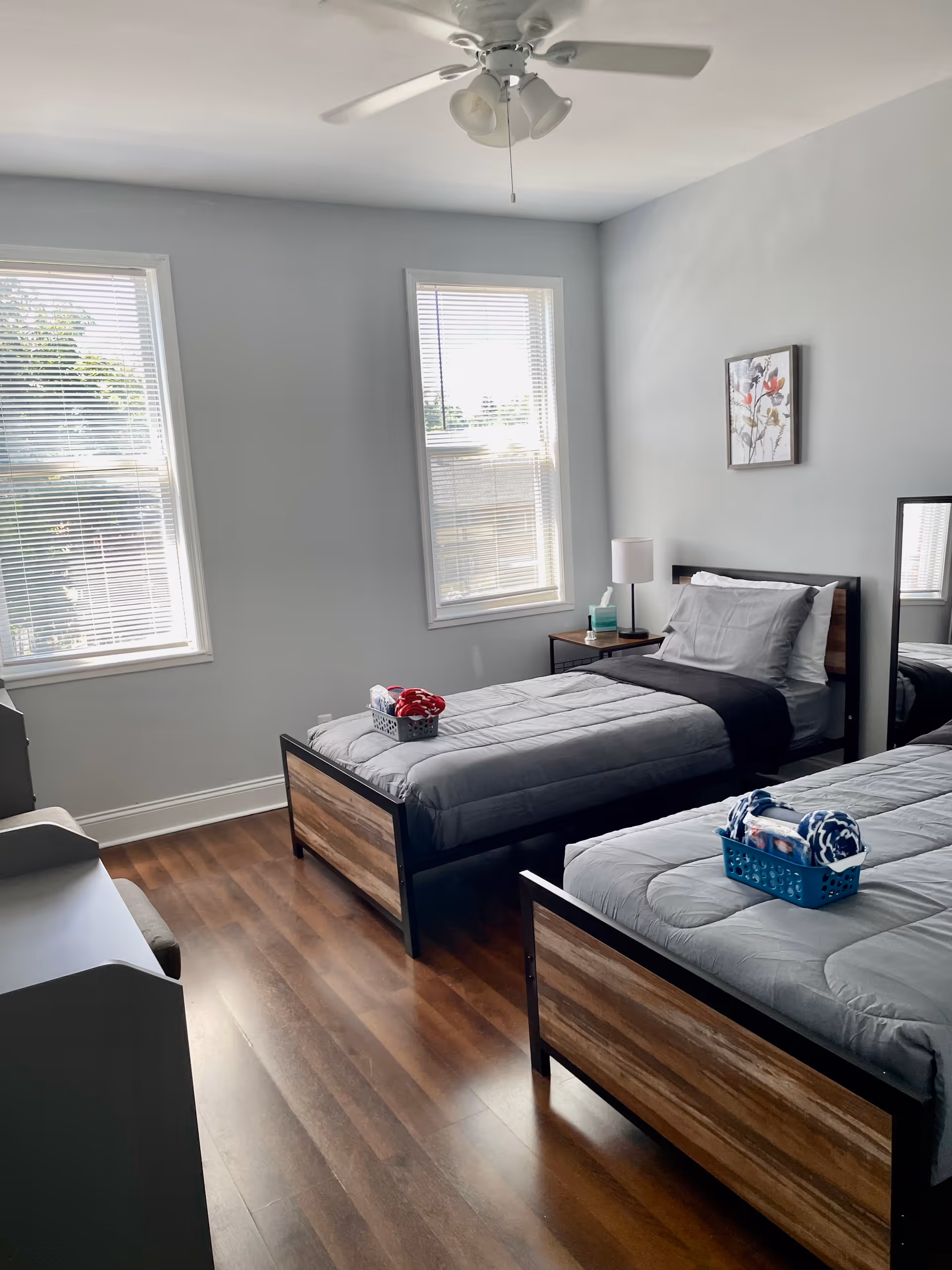 A bright bedroom with two single beds featuring wooden and metal frames, each bed made with gray bedding and a small basket of towels placed on top. The room has two tall windows with blinds, a wooden floor, a small nightstand with a lamp and tissues, a ceiling fan with lights, and a framed floral picture on the wall.