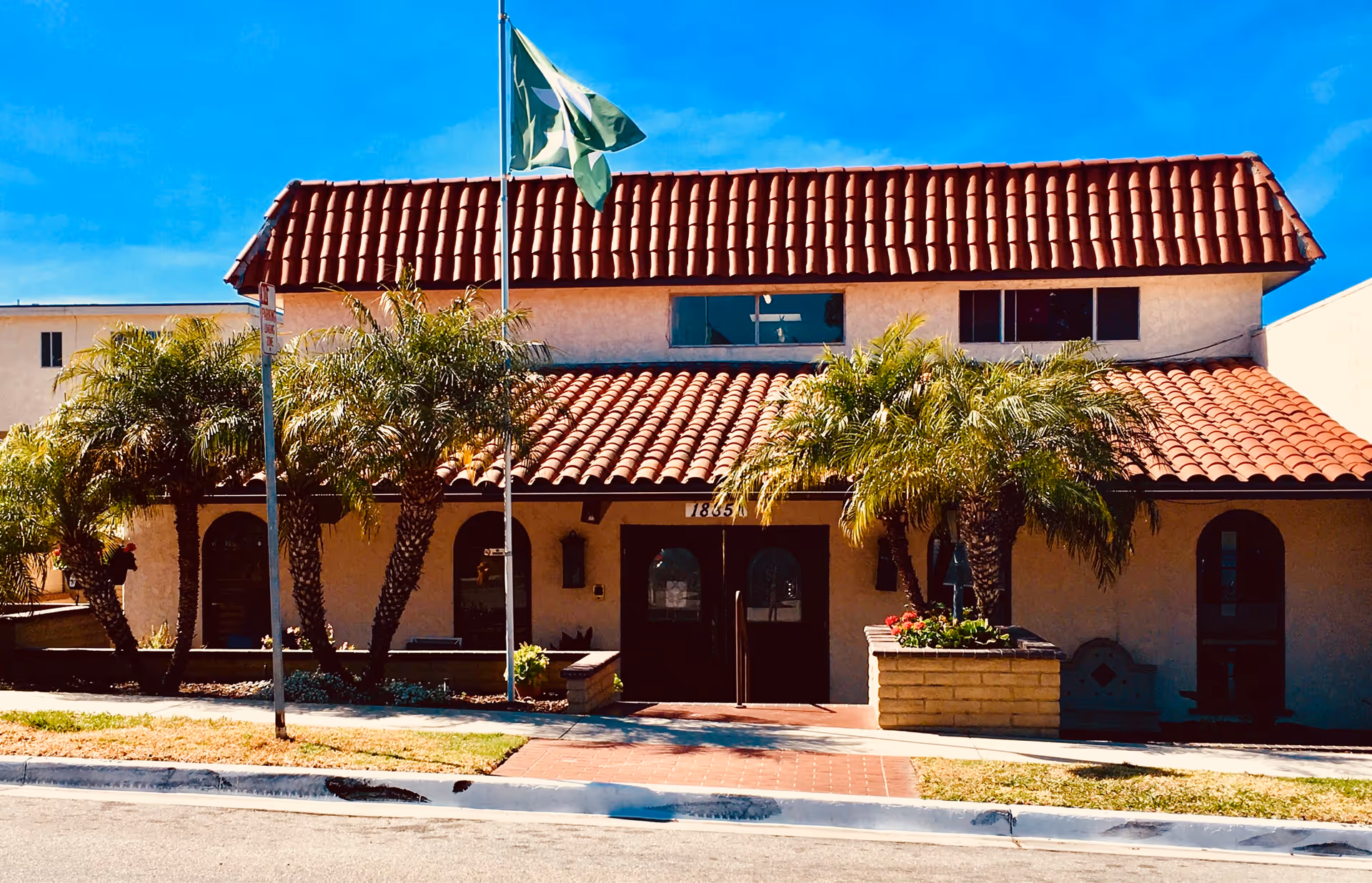 Exterior view of Sea Cliff Assisted Living facility with a red tiled roof, beige walls, and several palm trees in front. A flagpole with a green flag is positioned near the entrance, which has double doors and the number 1855 above them.