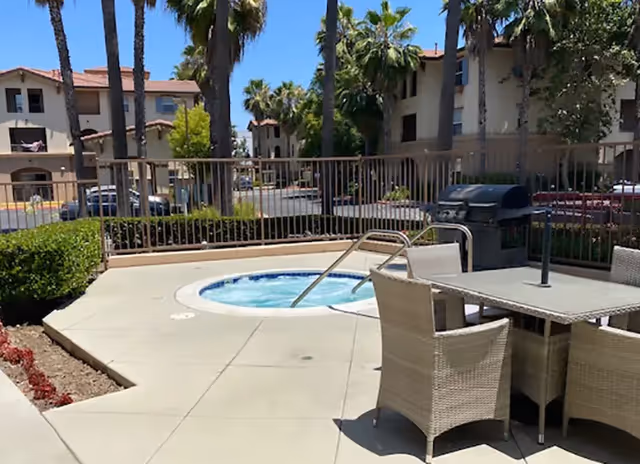 Outdoor patio area at Serenity Senior Villas featuring a round hot tub surrounded by a safety railing, a table with four wicker chairs, a barbecue grill, palm trees, and multi-story residential buildings in the background under a clear blue sky.