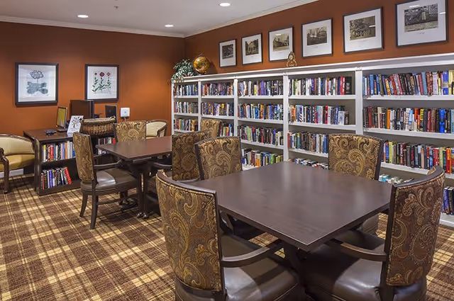 A cozy library room with two dark wood tables surrounded by patterned upholstered chairs. The walls are painted a warm brown, and there are white bookshelves filled with books along one wall. Above the bookshelves are framed black and white photographs. The floor is covered with a plaid carpet in shades of brown and beige. Additional seating and framed artwork are visible on the left side of the room.