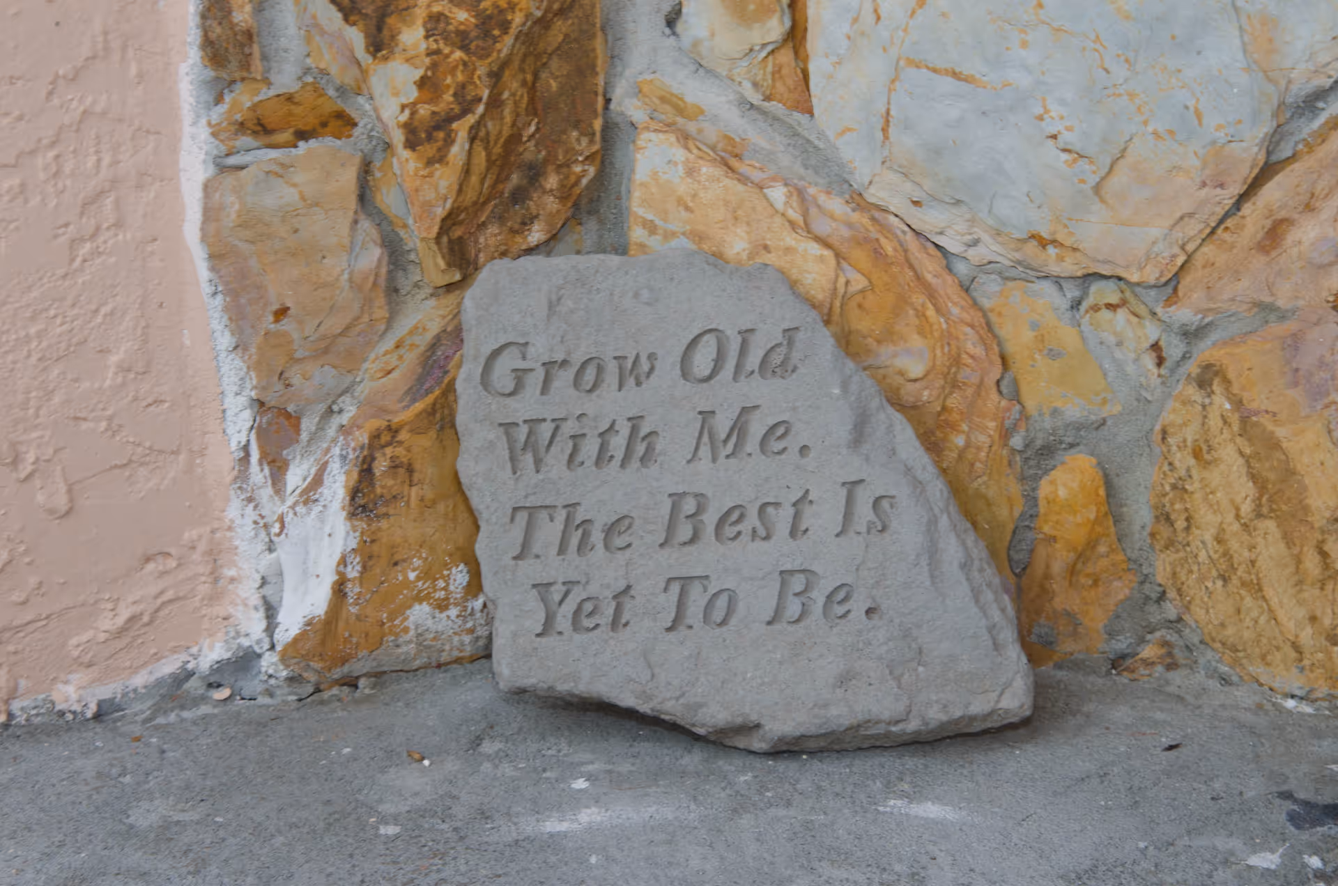 A stone plaque resting against a stone wall with the engraved inspirational message: 'Grow Old With Me. The Best Is Yet To Be.'