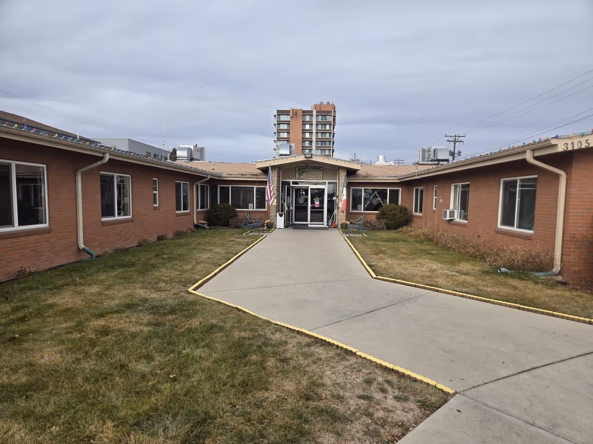 Front entrance of a single-story brick care facility with a central glass doorway, a paved walkway, and lawns on either side.