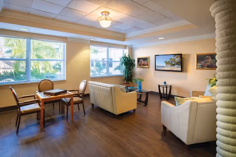 Bright communal sitting room with sofas, armchairs, a table, wall-mounted TV, and large windows overlooking greenery.