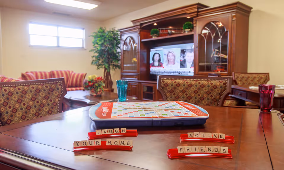A cozy living room area in a senior living facility with a wooden table in the foreground holding a Scrabble game board and letter racks spelling out 'LAUGH', 'YOUR HOME', 'ACTIVE', and 'FRIENDS'. In the background, there is a wooden entertainment center with a TV displaying a news program, a striped sofa, a potted plant, and patterned chairs around the table.