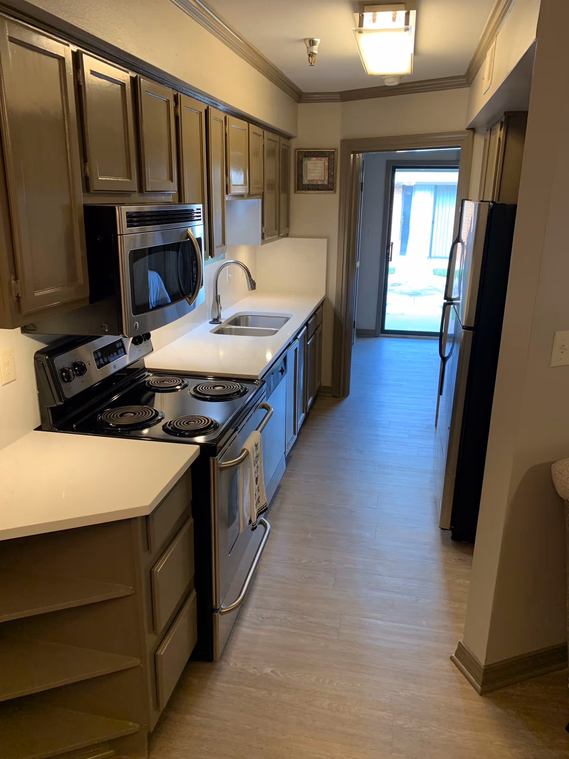 A narrow kitchen with wooden cabinets, a white countertop, a stainless steel sink, an electric stove with four coil burners, a microwave above the stove, a dishwasher, and a refrigerator. The kitchen has light wood flooring and a door at the end leading to an outdoor area.