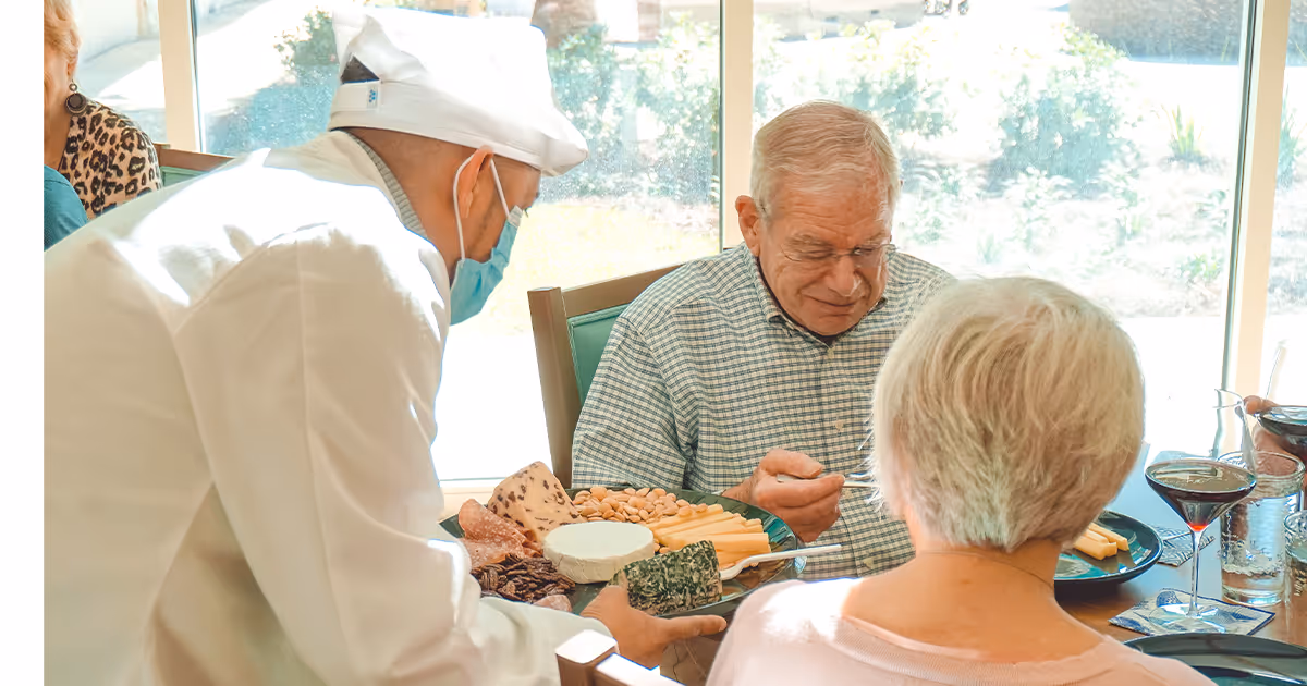 A masked chef serves a cheese and snack platter to two elderly residents seated at a dining table by a sunny window.