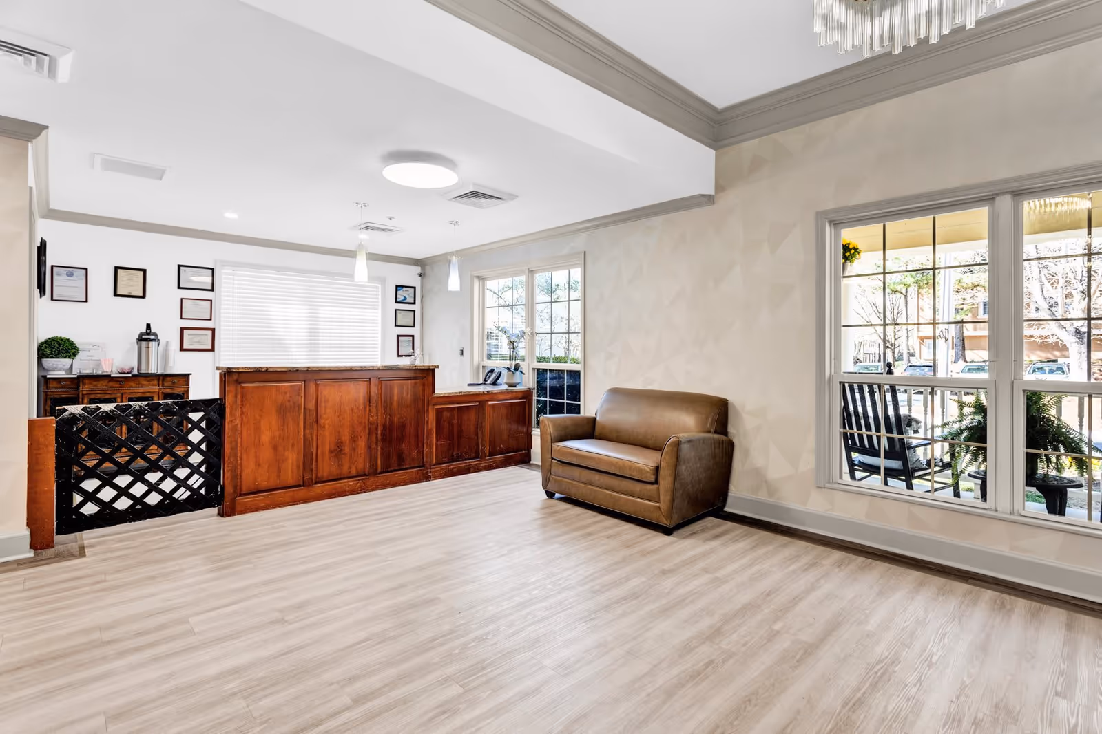 A bright and clean reception area of an assisted living facility featuring a wooden reception desk with certificates on the wall behind it, a small brown leather loveseat, large windows showing an outdoor porch with rocking chairs and plants, and light wood flooring.