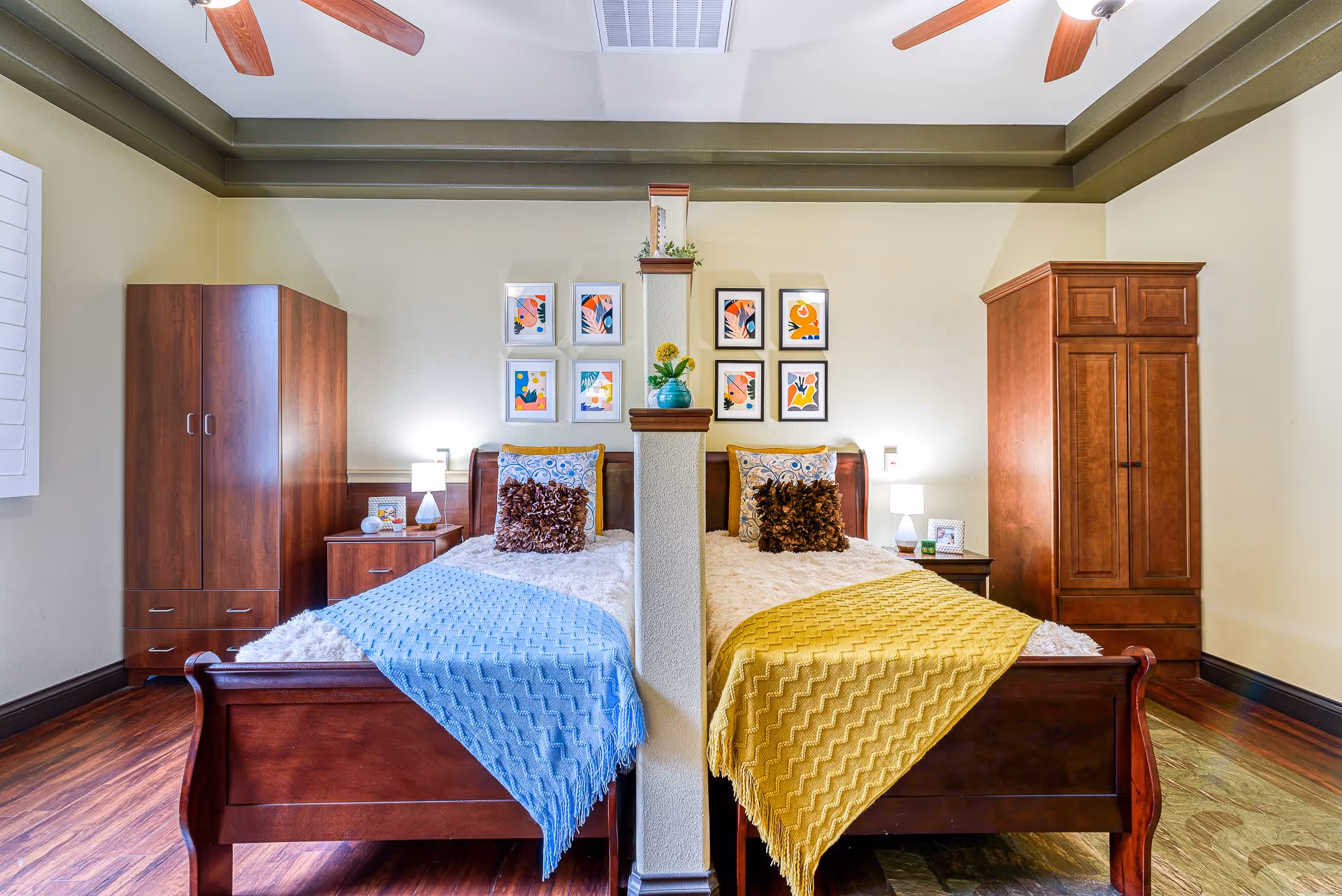 A bright bedroom with two wooden twin beds separated by a partial divider, colorful throws and pillows, nightstands, armoires, and framed artwork on the wall.