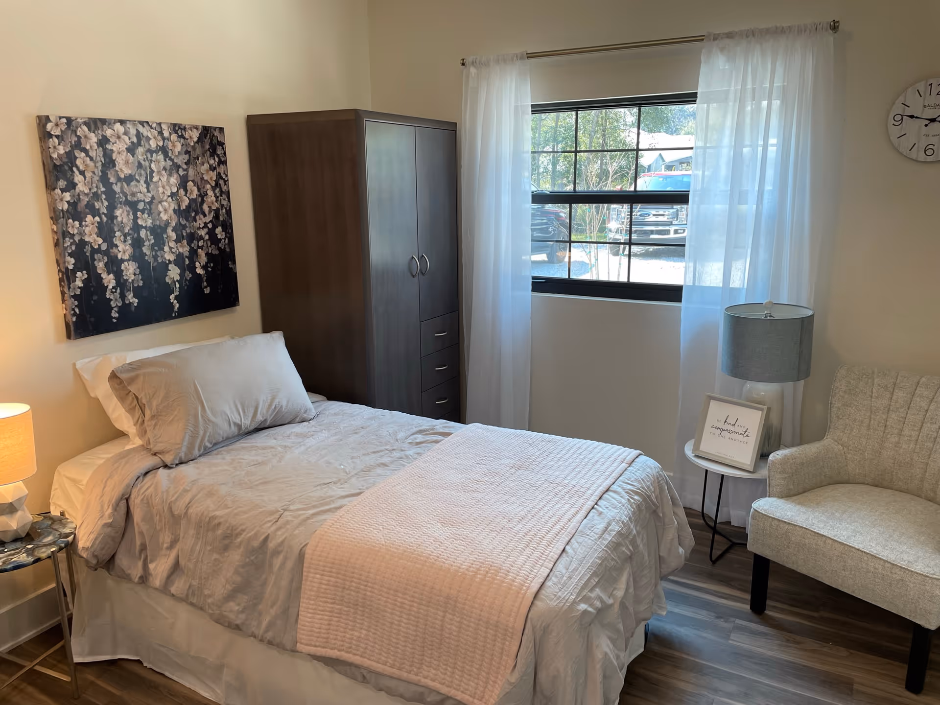 A cozy bedroom with a single bed made with white and light gray bedding. There is a dark wooden wardrobe next to the bed, a small round side table with a lamp, and a light gray upholstered chair. A window with sheer white curtains lets in natural light, and a floral artwork hangs on the wall above the bed. A clock is mounted on the wall near the window.