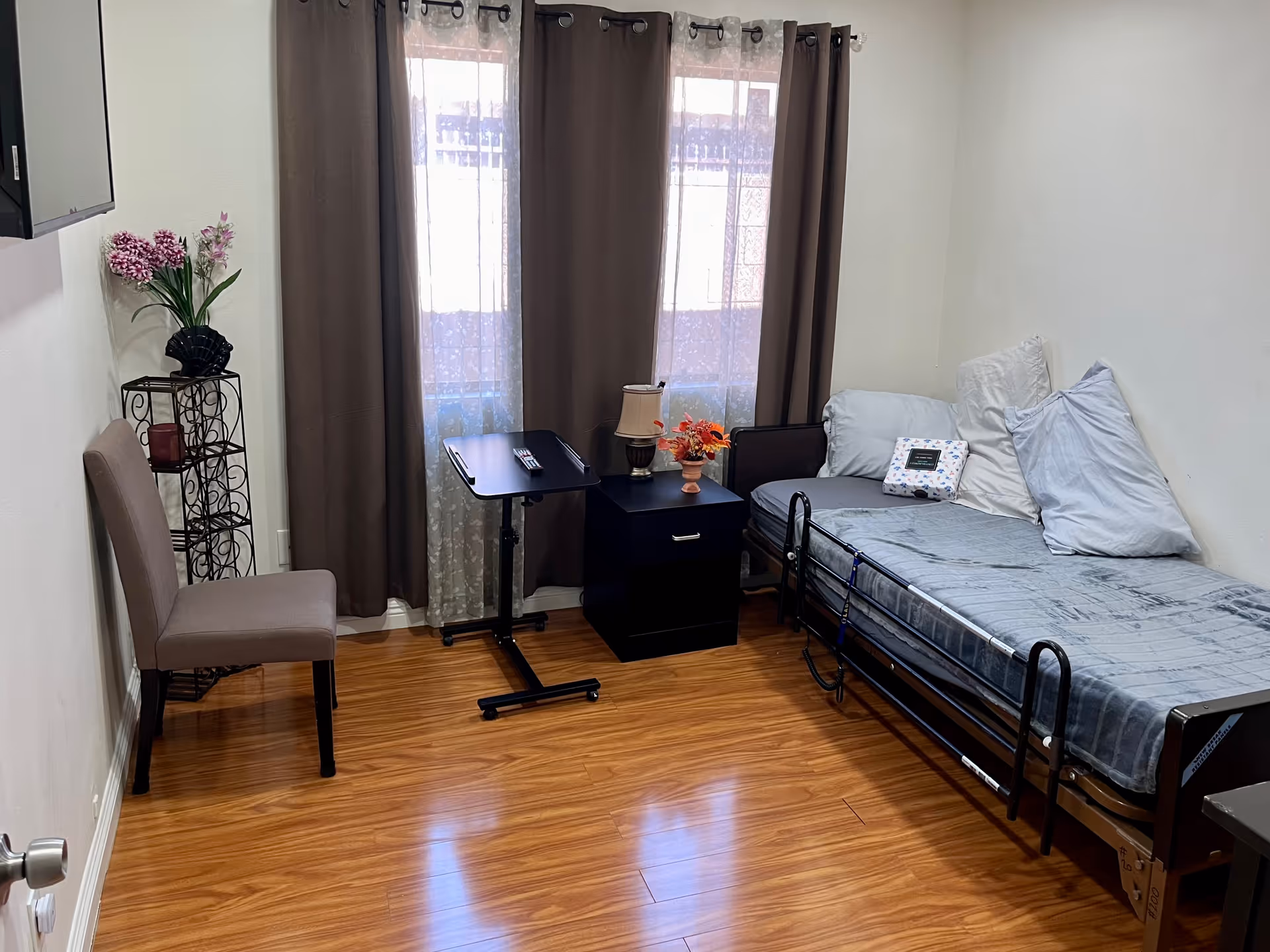 A small, tidy room with a single bed covered in gray bedding and multiple pillows against the right wall. Next to the bed is a black nightstand with a lamp and a small flower arrangement. In front of the window with brown curtains is a small black adjustable table. A beige upholstered chair is placed against the left wall near a decorative metal stand holding a vase with pink flowers. The floor is wooden and the walls are white.