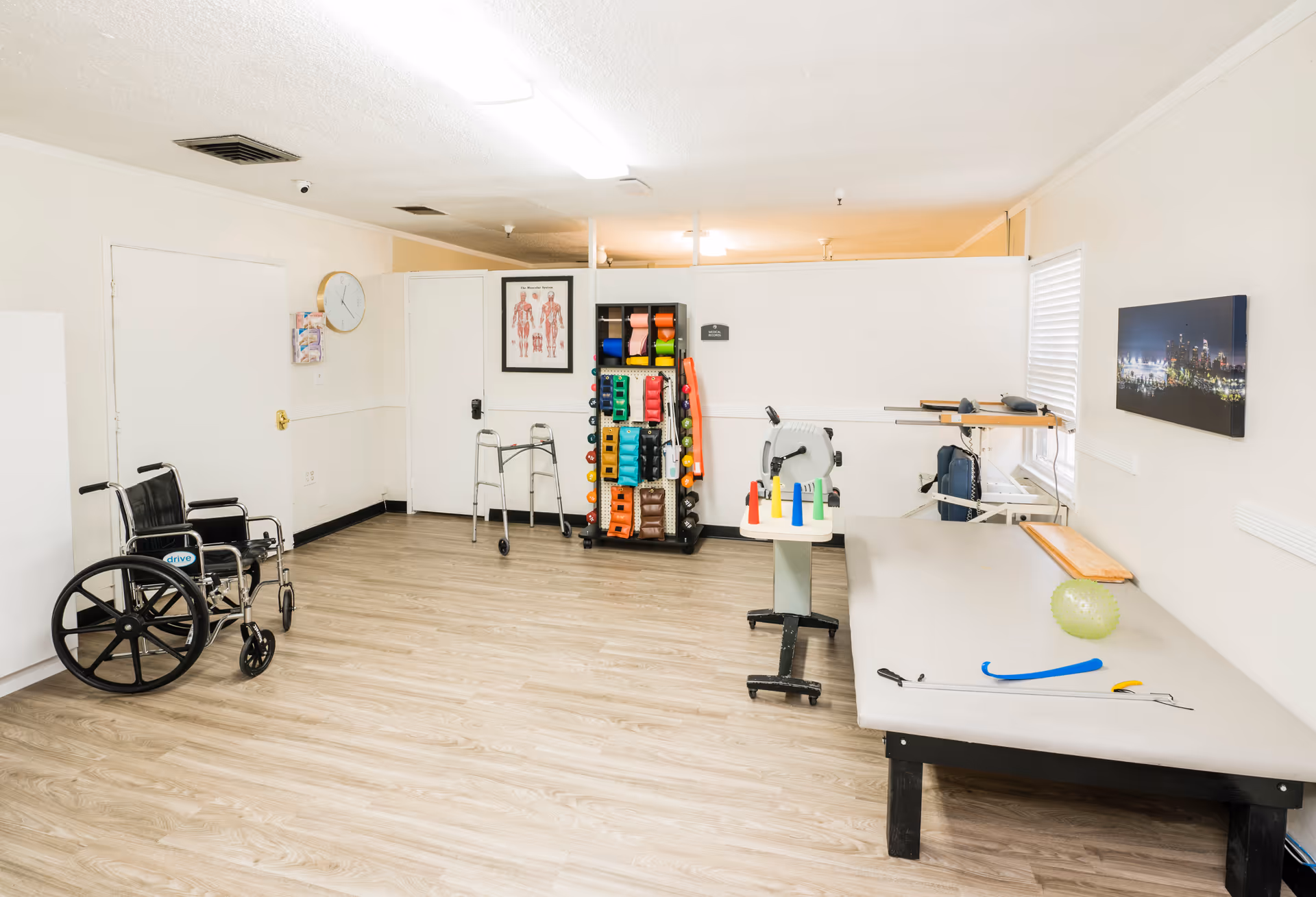 A rehabilitation room with a wheelchair, walker, exercise equipment, therapy table with various therapy tools, and anatomical chart on the wall. The room has light wood flooring, white walls, and a window with blinds.