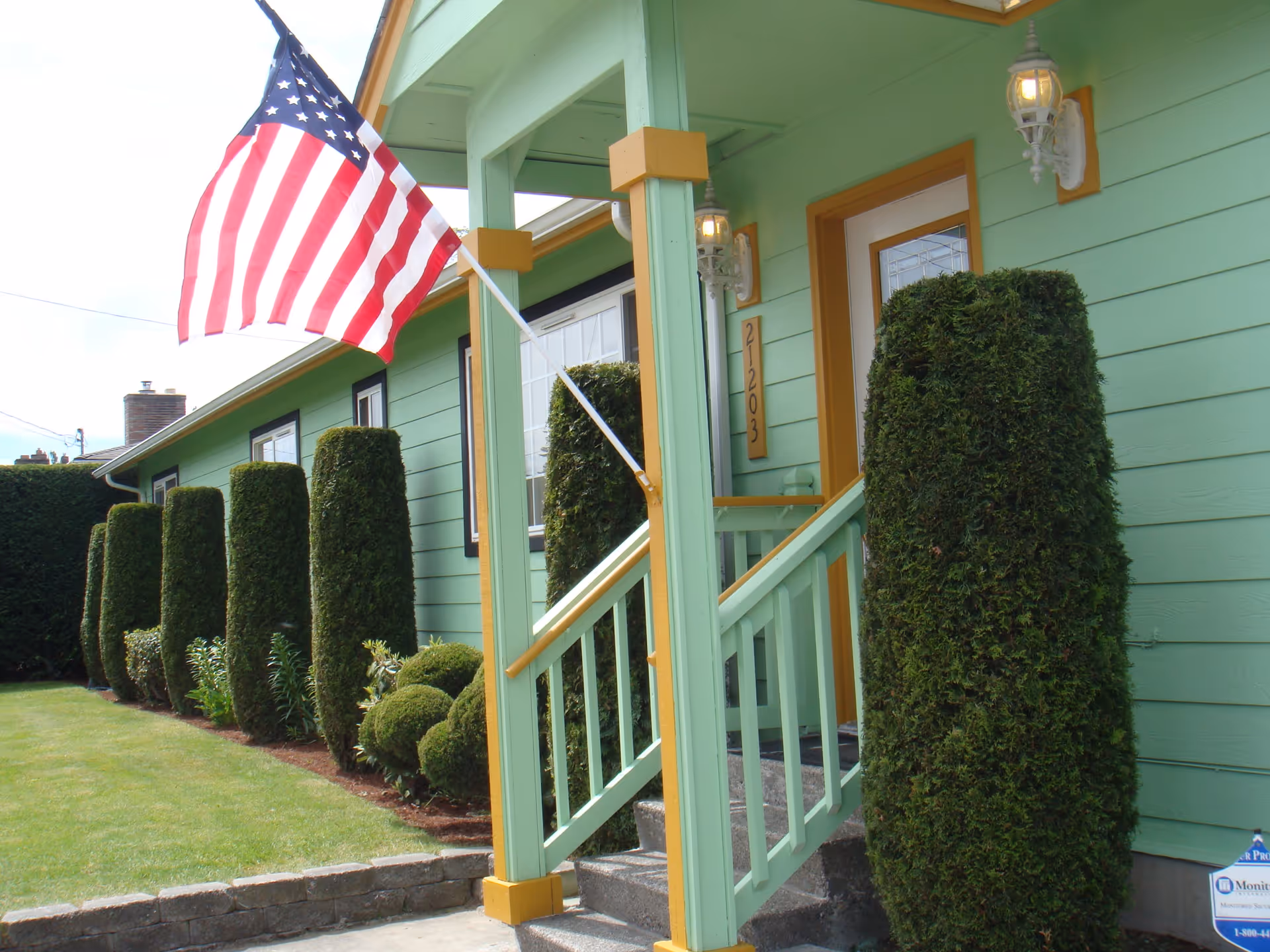 Front porch of a green house with yellow trim, featuring neatly trimmed tall bushes along the side and an American flag mounted on a pole near the entrance. The house number 21203 is visible next to the door.