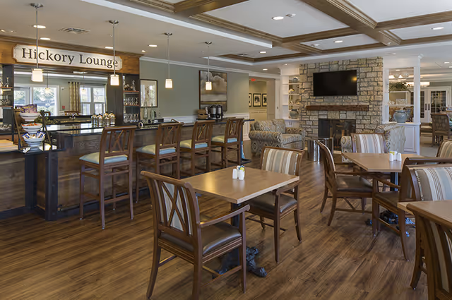 Interior view of the Hickory Lounge at Brightview Tenafly, featuring a bar area with high chairs, wooden tables and chairs, a stone fireplace with a mounted TV above it, and comfortable armchairs. The room has wooden flooring, pendant lights, and a cozy, inviting atmosphere.