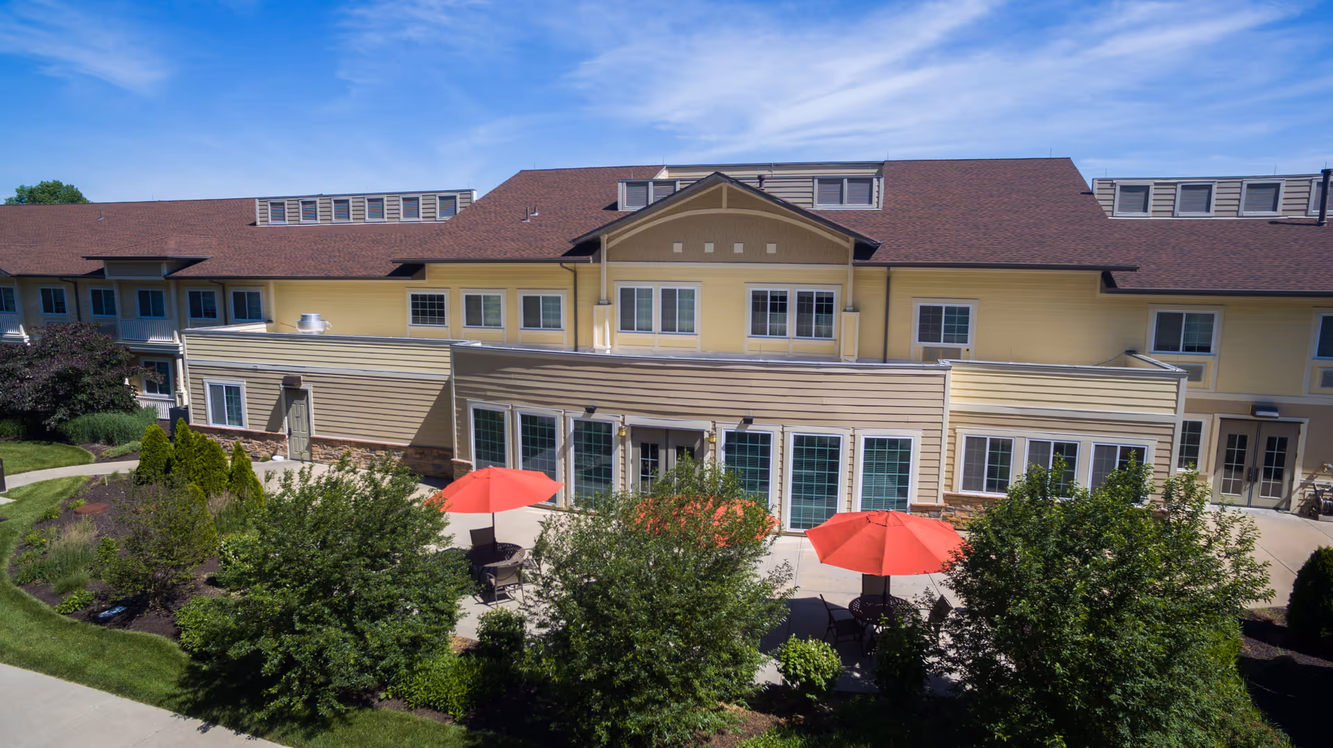 Exterior view of Westview at Ellisville Assisted Living facility showing a two-story building with beige siding and a brown roof. There is a patio area with red umbrellas and outdoor seating surrounded by green bushes and landscaping under a clear blue sky.