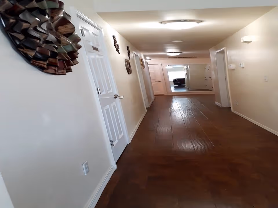 A long hallway with wooden flooring and cream-colored walls. Several white doors line the hallway on both sides. Decorative wall hangings are visible on the left wall, and a large mirror is mounted on the far end wall reflecting part of a room with a couch. The hallway is well-lit with ceiling lights.