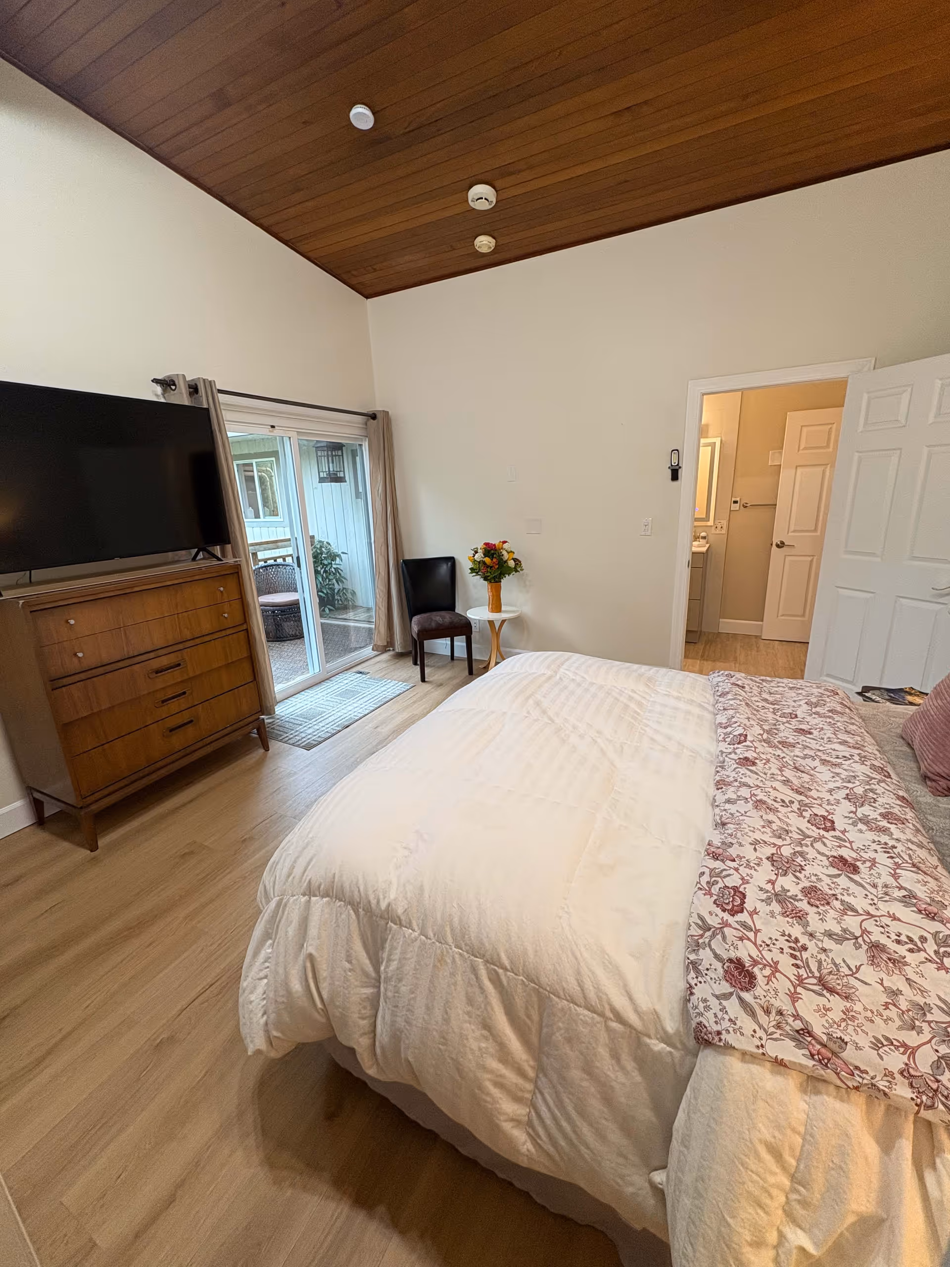 Bright bedroom with a bed, wooden dresser and TV, sliding glass door to a patio, and a doorway leading to a bathroom.