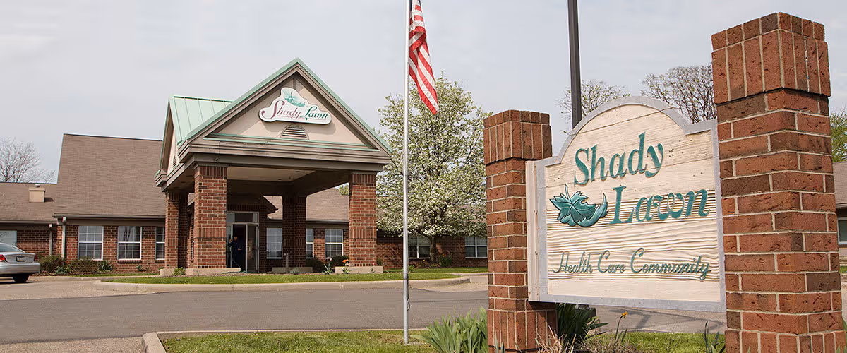 Exterior view of The Oaks at Shady Lawn health care community building with a brick entrance and a sign displaying the facility name. An American flag is visible near the entrance.