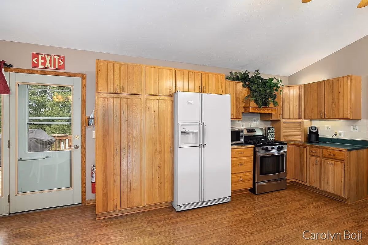 A kitchen with wooden cabinets, a white side-by-side refrigerator, a stainless steel gas stove, a microwave, a coffee maker, and a green countertop. There is a door with a window leading outside, above which is a red EXIT sign. A plant is placed on top of the cabinets above the stove.