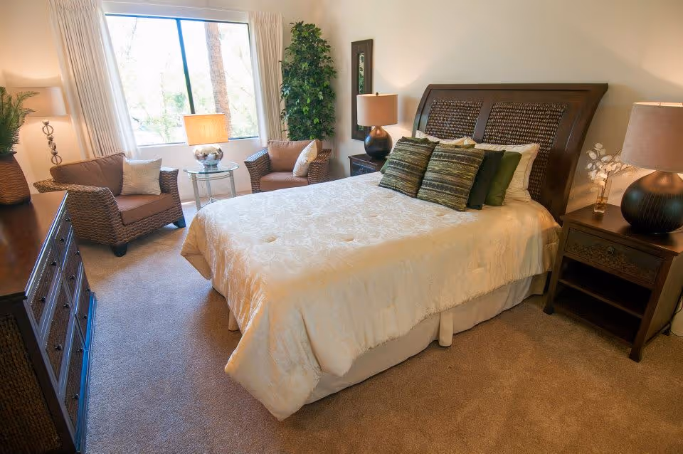 A cozy bedroom with a large bed covered in a cream-colored comforter and several green and patterned pillows. There are two wooden nightstands with lamps on either side of the bed. To the left, there are two wicker armchairs with cushions and a small round glass table with a lamp between them. A large window with white curtains lets in natural light, and a tall green plant is placed in the corner near the window. A wooden dresser is visible on the left side of the room.