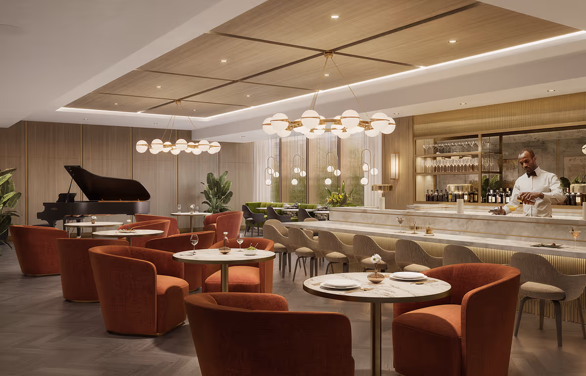 Stylish communal dining and lounge area with round marble tables, red upholstered chairs, a marble bar with a bartender, and a grand piano in the background.