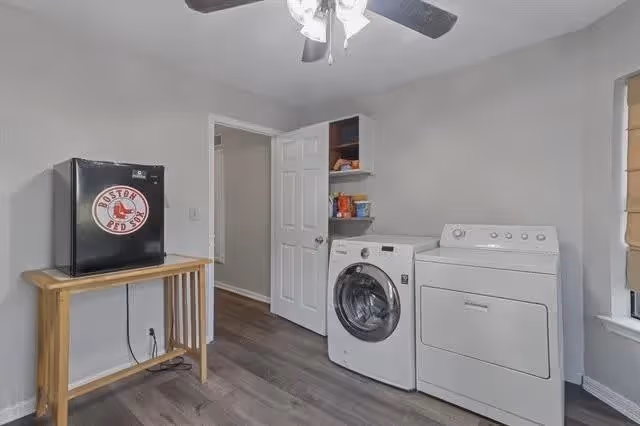 A laundry room with a front-loading washing machine and a top-loading dryer side by side. There is a small wooden table with a black mini fridge on top, which has a Boston Red Sox logo sticker. The room has light gray walls, a ceiling fan with lights, and a window with a beige blind partially visible on the right.