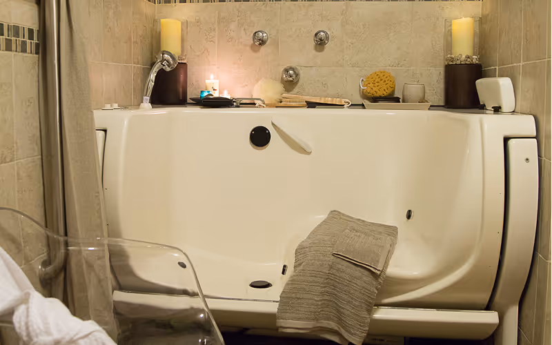 A walk-in bathtub in a bathroom with beige tiled walls. The tub has a towel draped over the side, a handheld showerhead, and various bath accessories including candles, a sponge, and a brush placed on the tub's edge. A transparent chair is partially visible in front of the tub.