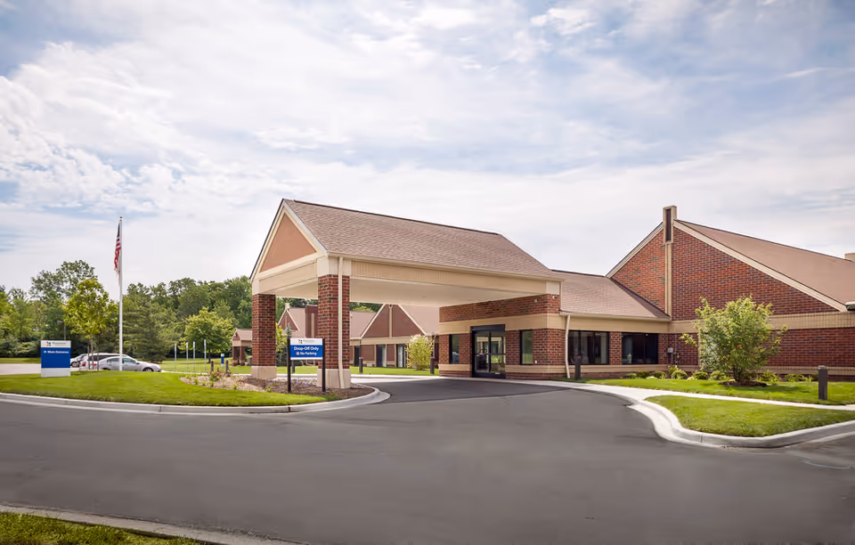 Front exterior view of Pomeroy Living Sterling Skilled Rehabilitation building with a covered drop-off area, brick walls, and a paved driveway surrounded by green lawns and trees under a partly cloudy sky.