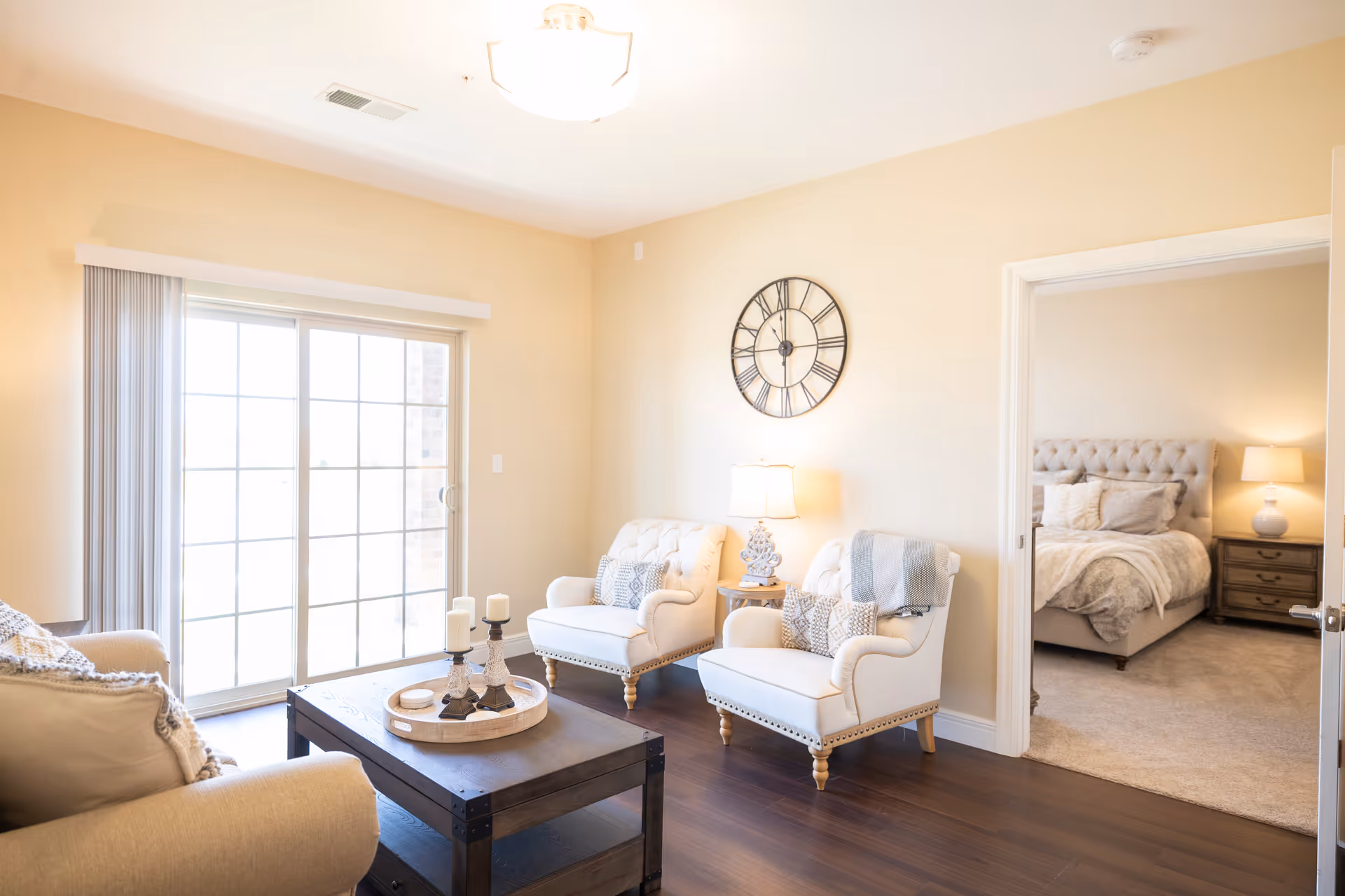A bright and cozy living room with two white armchairs, a beige sofa, a wooden coffee table with candles, and a large wall clock. Sliding glass doors allow natural light to fill the room. An adjacent bedroom with a tufted bed and nightstand with a lamp is visible through an open doorway.