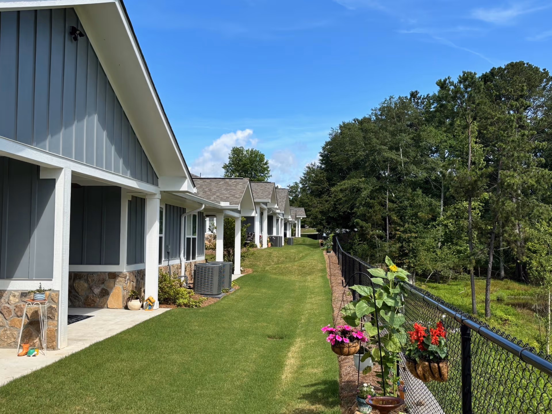 A row of single-story residential buildings with gray siding and stone accents, each with a small covered porch. A well-maintained grassy area runs alongside the buildings, bordered by a black chain-link fence with hanging flower pots containing colorful flowers. Trees and greenery are visible beyond the fence under a clear blue sky.
