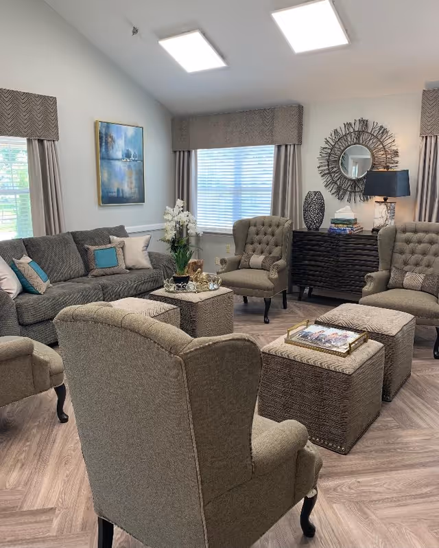 Bright senior living room with upholstered sofas and armchairs arranged around ottomans and a coffee table, with a decorative mirror and sideboard.