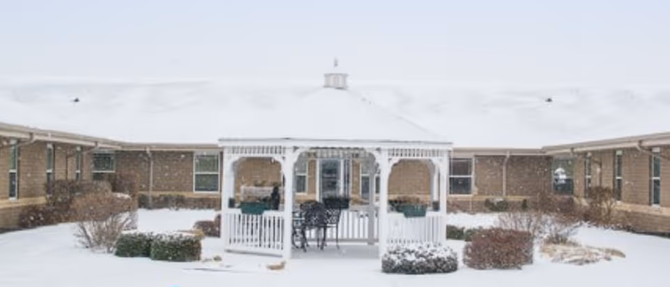 Snow-covered courtyard of a retirement community featuring a white gazebo with chairs and tables inside, surrounded by a single-story brick building with multiple windows.