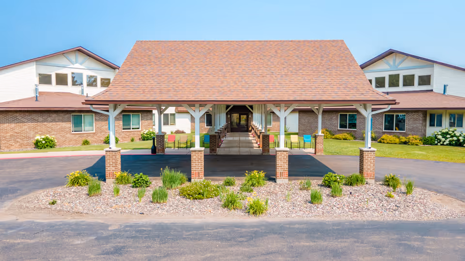 Front exterior view of a senior living facility named Gardenview featuring a covered entrance with brick pillars, a paved driveway, landscaped plants, and a building with brick and white siding under a clear blue sky.