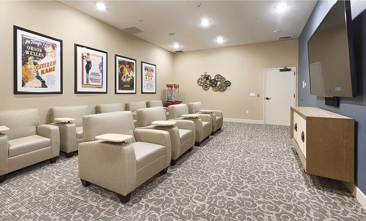 A small theater room with two rows of beige armchairs, each equipped with a small wooden tray. The walls are decorated with framed classic movie posters and a decorative wall piece made of film reels. A large flat-screen TV is mounted on the wall opposite the chairs, above a wooden cabinet. The room has patterned carpet flooring and recessed ceiling lights.