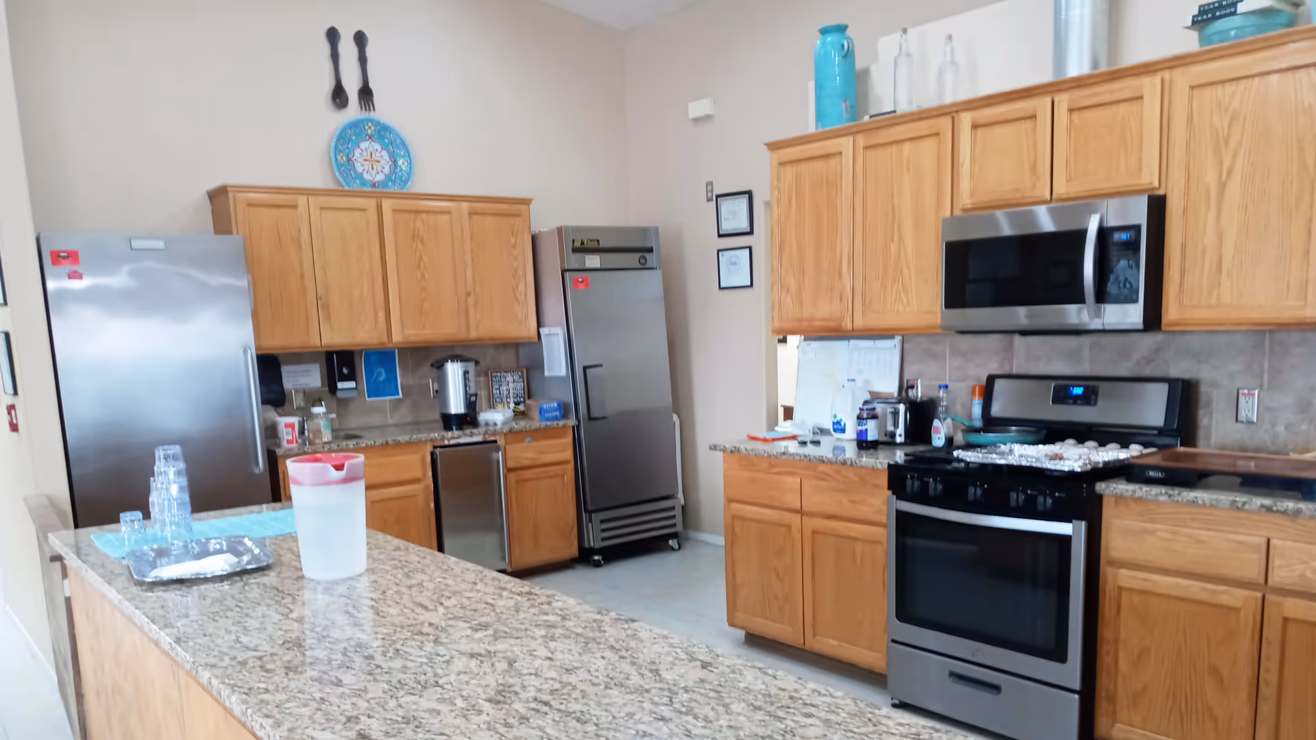 A clean, well-lit kitchen with wooden cabinets, granite countertops, a stainless steel refrigerator, a microwave, and a stove with a tray of muffins. There is a water pitcher and stacked plastic cups on the kitchen island in the foreground.