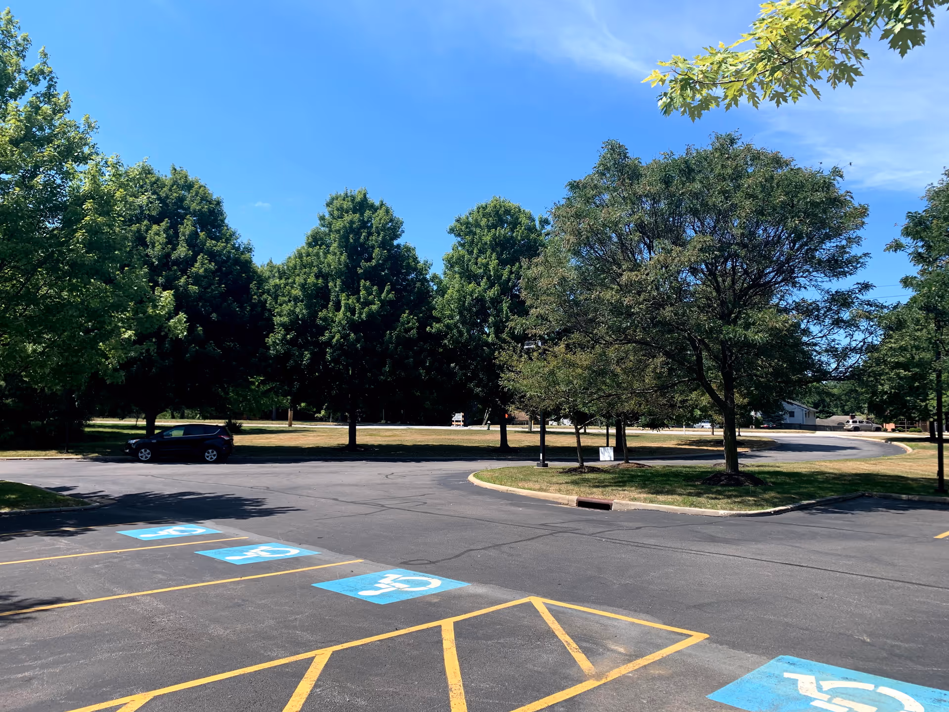 A parking lot with marked handicap spaces, a parked car, trees and a grassy area under a clear blue sky.