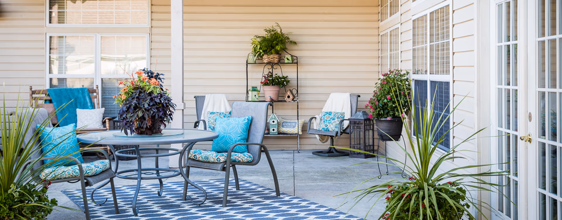 Outdoor patio area with a round glass table surrounded by four cushioned chairs with blue and patterned pillows. There are various potted plants and flowers around the patio, including tall green plants in the foreground and colorful flowers on a metal shelf against the beige siding wall. Two rocking chairs with cushions and a small side table are also visible near the windows and glass doors.