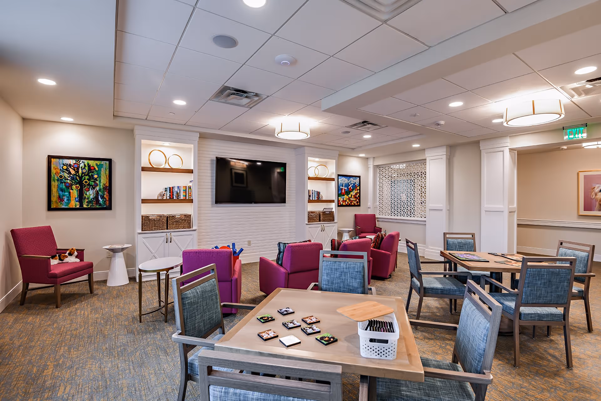 A well-lit common area in a senior living facility featuring several tables with chairs, a group of purple armchairs arranged around a large flat-screen TV mounted on the wall, decorative shelving with books and baskets, colorful artwork on the walls, and a patterned carpeted floor.