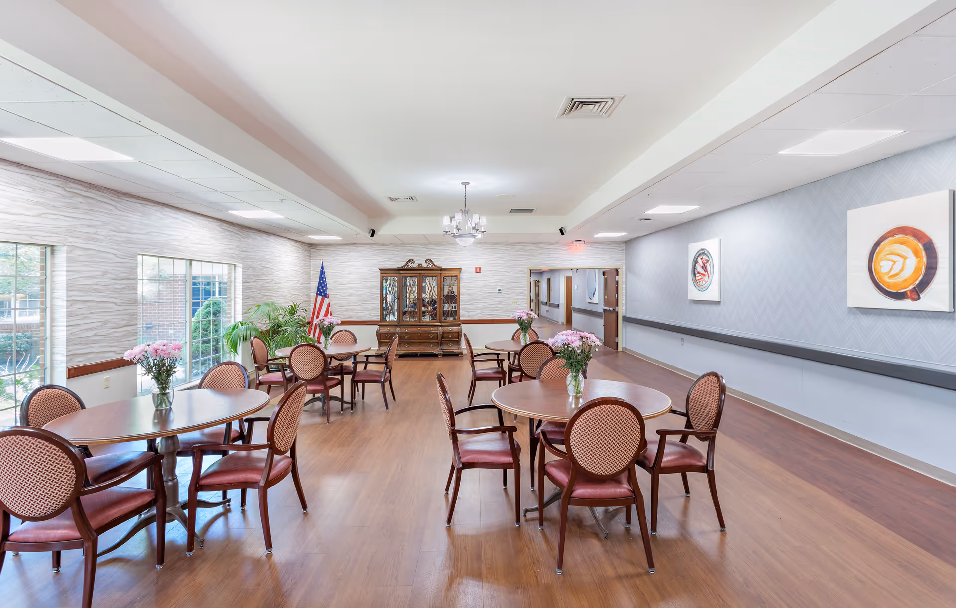 A bright and spacious dining room with round wooden tables and cushioned chairs arranged neatly. Each table has a vase with pink flowers. Large windows on the left side let in natural light, and there is an American flag and a wooden cabinet at the far end of the room. The walls are decorated with framed artwork, and the floor is wooden.