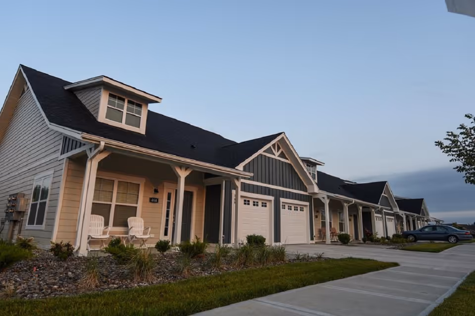Front exterior of a row of single-story attached homes with porches, garages, and a sidewalk.