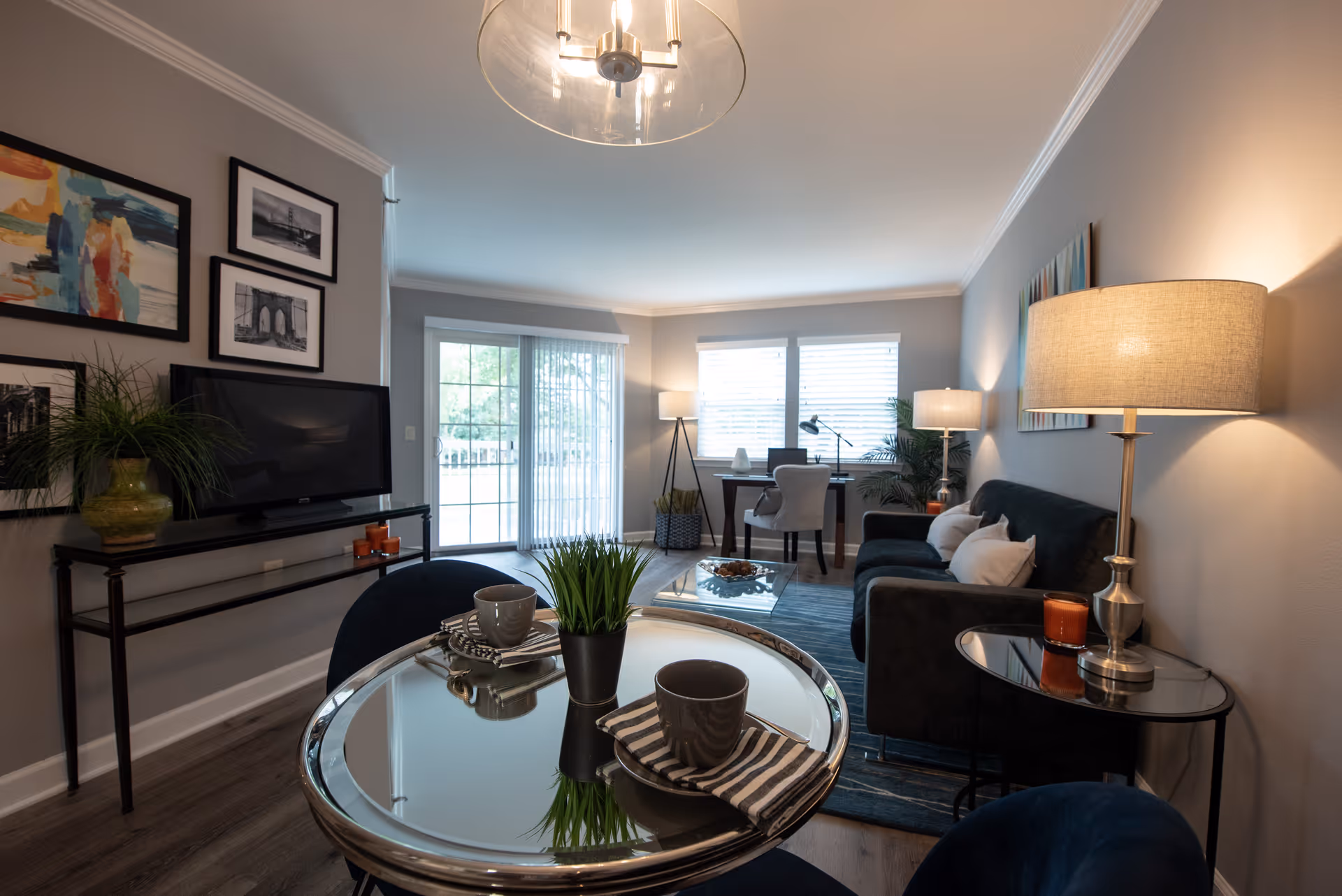 A cozy living room area with a small round glass table set with two cups and napkins, a dark sofa with cushions, a side table with a lamp and candle, a TV on a slim black stand, framed artwork on the wall, and a desk with a chair and lamp near windows with blinds and a sliding glass door leading outside.