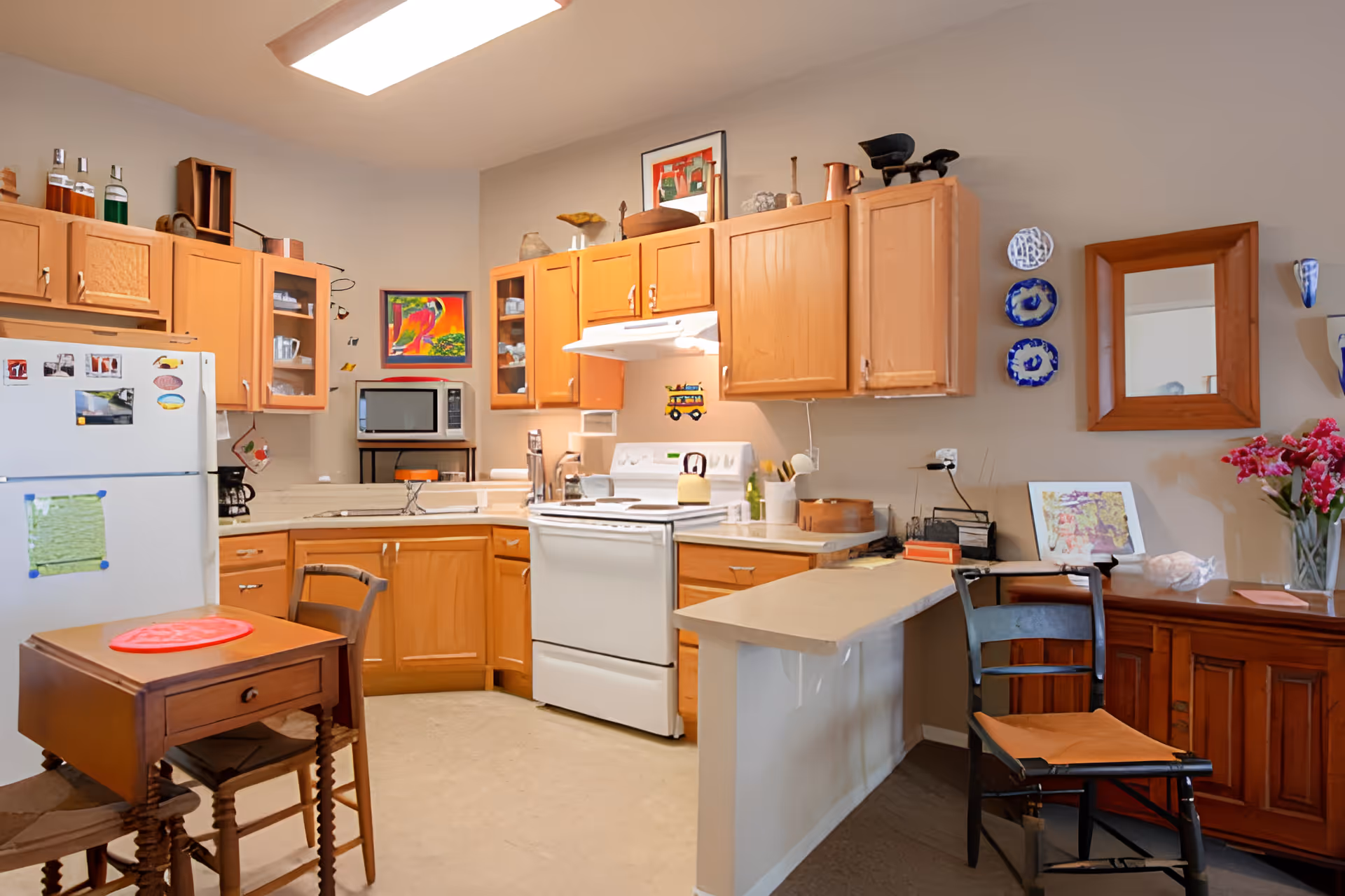 Well-lit kitchen with wooden cabinets, white appliances, a small table and chairs, and decorative plates on the wall.