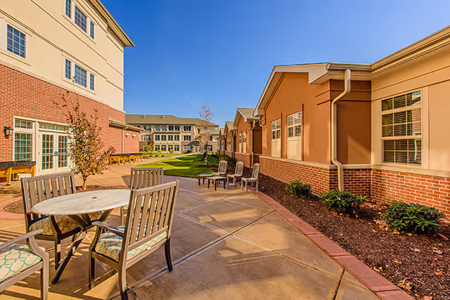 Outdoor patio area at Brookdale Gables Farmington with round tables and chairs on a paved walkway, surrounded by buildings with brick and beige siding under a clear blue sky.