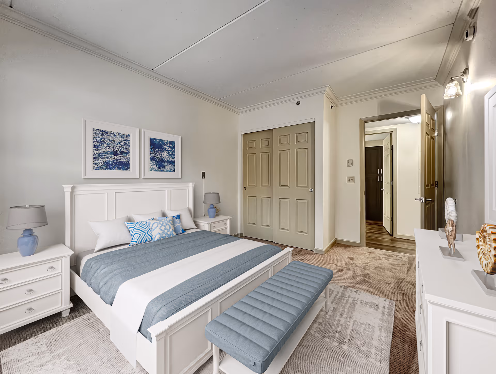 A neatly arranged bedroom with a white bed frame and headboard, blue and white bedding, and a matching blue bench at the foot of the bed. There are two white nightstands with blue lamps on either side of the bed. Two framed pictures of ocean waves hang above the headboard. The room has beige carpet, a white dresser with decorative items on top, and a closet with double doors. An open door leads to a hallway with wood flooring.