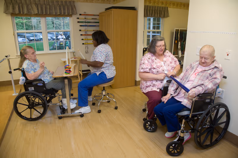 Two elderly women in wheelchairs engaged in activities with two caregivers in a bright room with wooden flooring and windows showing parked cars outside. One caregiver is seated on a stool playing a game with one woman, while the other caregiver is assisting another woman with a tablet device.