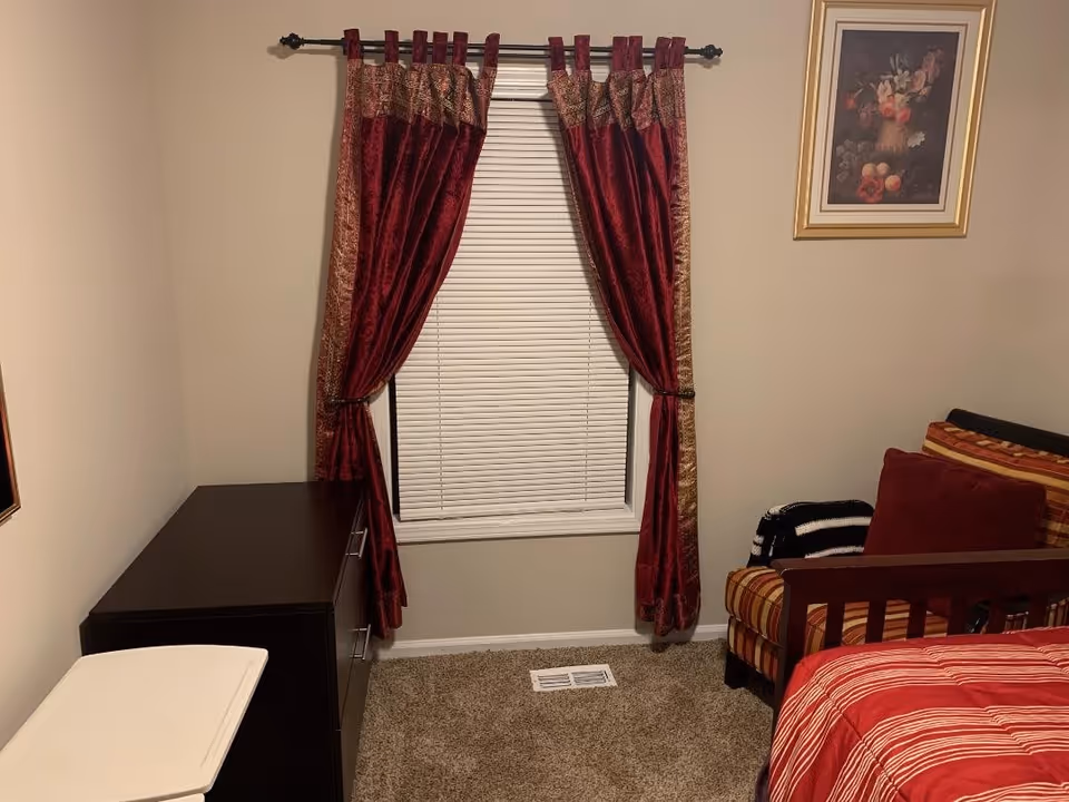 A small bedroom with beige walls and carpeted floor. There is a window with closed white blinds and red curtains with gold trim. To the left of the window is a dark brown dresser, and to the right is a striped armchair with a red pillow and a black and white blanket. Part of a bed with a red and white striped comforter is visible in the foreground. A framed floral painting hangs on the wall above the armchair.