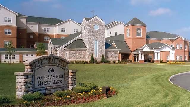 Exterior view of Mother Angeline McCrory Manor, a large senior living facility with a stone and brick facade, green roofs, and a well-maintained lawn with a stone sign displaying the facility's name in the foreground.