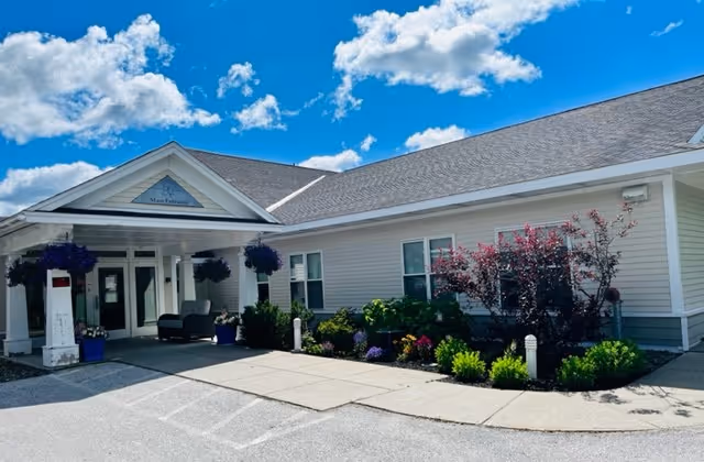 Exterior view of a single-story senior living facility building with a covered entrance, hanging flower baskets, and landscaped bushes and flowers under a bright blue sky with scattered clouds.