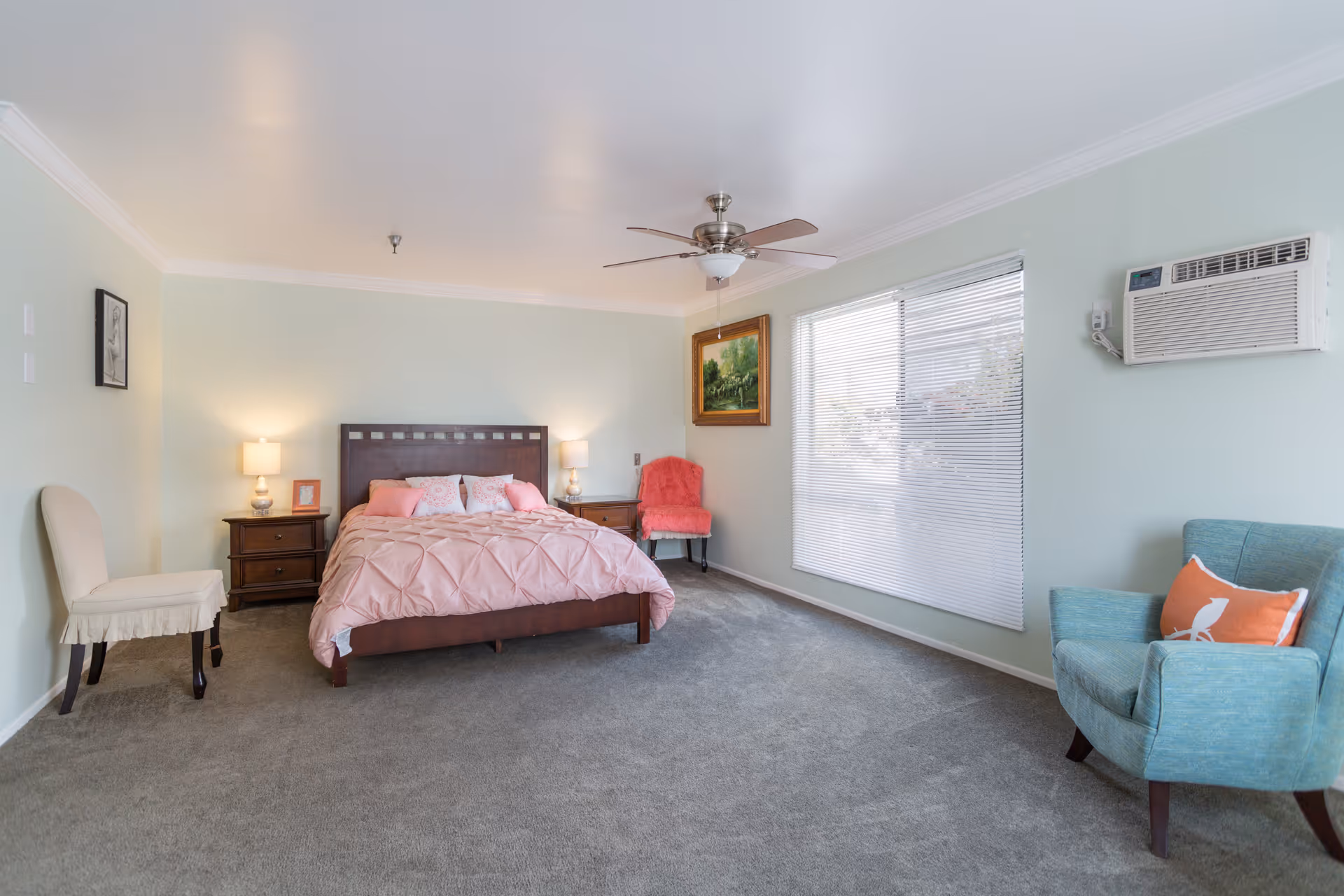 A spacious bedroom with a large bed covered in a pink comforter and pillows. There are two wooden nightstands with lamps on either side of the bed. The room has light green walls, a ceiling fan, a large window with blinds, and three chairs: one beige, one pink, and one blue with an orange pillow. A framed painting hangs on the wall near the window, and an air conditioning unit is mounted on the wall.