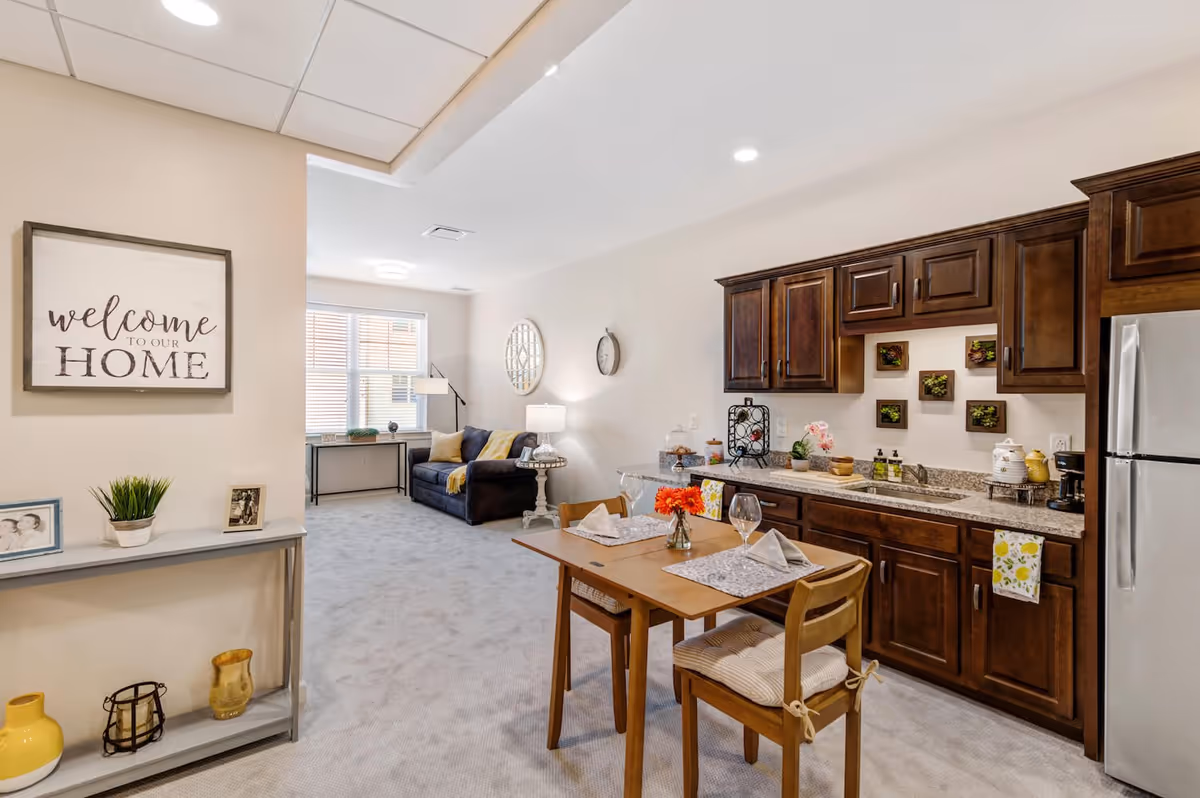 A cozy assisted living apartment interior featuring a small wooden dining table set for two with placemats, napkins, and a vase of orange flowers. To the right is a kitchenette with dark wooden cabinets, a granite countertop, a stainless steel refrigerator, and various kitchen items. In the background, there is a living area with a dark sofa, a side table with a lamp, a wall clock, and a decorative mirror. On the left wall, a framed sign reads 'welcome to our HOME' above a small shelf holding framed photos, a plant, and decorative items.